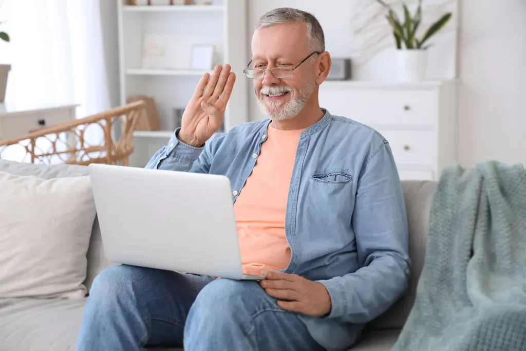 Middle aged man waving at camera on laptop while sitting on couch